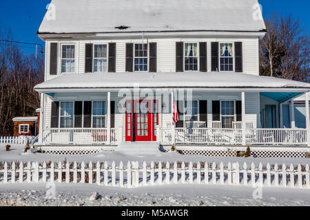 Un tradizionale dipinto di bianco Englander Nuova casa con un wrap-around portico, rosso porta anteriore nero, persiane, bandiera americana, Picket Fence e un cortile pieno o Foto Stock