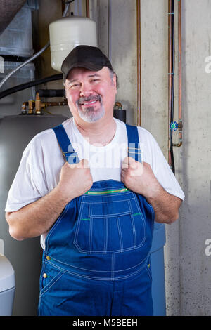 Trustworthy middle-class operaio in un seminterrato sala di utilità in piedi tenendo la sua tuta in jeans come egli sorride alla telecamera Foto Stock