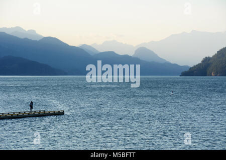 Una donna sta alla fine del dock sul lago Harrison, con la costa delle montagne sullo sfondo, in Harrison Hot Springs, British Columbia, Canada. Foto Stock