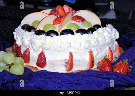 Torta guarnita con frutta. Torta alla frutta con panna, fette di mela, fragole, uve bianche uve nere. Close up di torta bianca coperta con frutti. Foto Stock