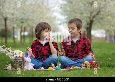 Due bambini, ragazzo fratelli, mangiando cioccolato coniglietti e divertirsi con le uova di pasqua nel parco, splendida fioritura primaverile giardino Foto Stock