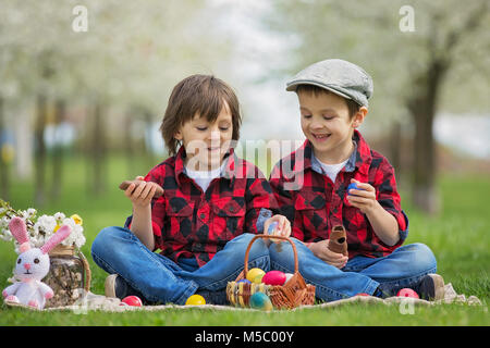 Due bambini, ragazzo fratelli, mangiando cioccolato coniglietti e divertirsi con le uova di pasqua nel parco, splendida fioritura primaverile giardino Foto Stock