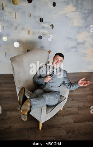 Bel uomo in abito grigio con microfono seduto sulla sedia studio dello sfondo. Toastmaster e mattatore. Foto Stock