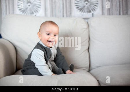 Little baby boy, seduto su un divano in un soleggiato soggiorno, sorridente felicemente Foto Stock