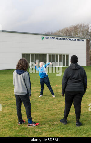 Gli studenti fanno pratica con la riproduzione di cricket su una scuola campo da gioco come parte di una lezione di giochi con il loro insegnante Foto Stock