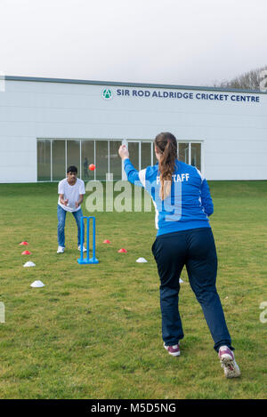 Gli studenti fanno pratica con la riproduzione di cricket su una scuola campo da gioco come parte di una lezione di giochi con il loro insegnante Foto Stock