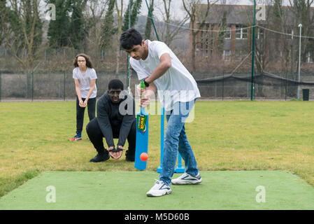 Gli studenti fanno pratica con la riproduzione di cricket su una scuola campo da gioco come parte di una lezione di giochi con il loro insegnante Foto Stock