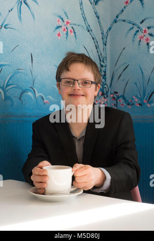 Un giovane contento e fiducioso uomo con la sindrome di down si siede a un tavolo in un caffè Foto Stock