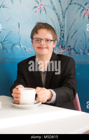 Un giovane uomo con la sindrome di down si siede a un tavolo in un bar a bere caffè Foto Stock