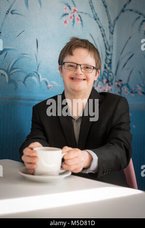 Un giovane uomo con la sindrome di down si siede a un tavolo in un bar a bere caffè Foto Stock