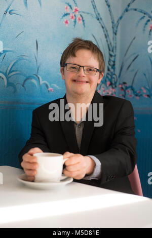 Un giovane uomo con la sindrome di down si siede a un tavolo in un bar a bere caffè Foto Stock