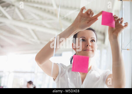 Bella giovane imprenditrice mettendo sticky notes sulla parete di vetro in office Foto Stock