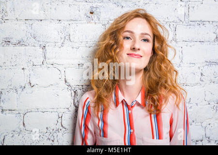 Ritratto di un bel giovane e bella donna studente con il rosso dei capelli ricci e le lentiggini sul suo viso è appoggiata contro un muro di mattoni di colore grigio. Dre Foto Stock
