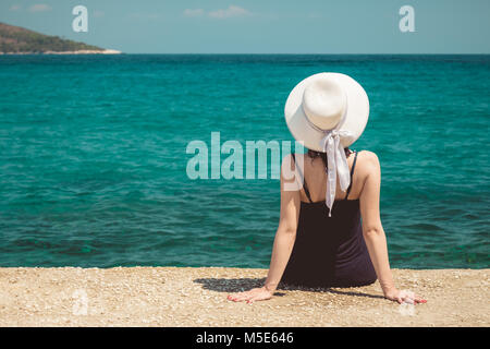 Vista posteriore della femmina elegante indossando cappello per il sole, seduto sul bordo del dock di calcestruzzo e godendo della vista sul mare. Viaggi e vacanze estive concetti. Foto Stock