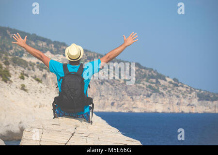 Vista posteriore del giovane turista ragazzo seduto sulla roccia con mani e godendo della vista sul mare Foto Stock