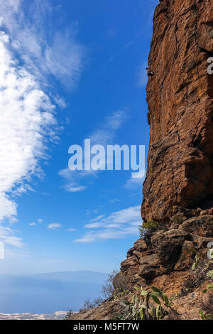 Maschio di rocciatore sulla ripida scogliera rossa con cactus e mare blu cielo blu dietro Foto Stock