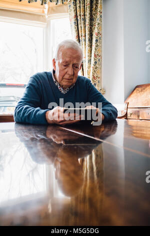 L'uomo anziano è seduto al tavolo della cucina nella sua casa, guardando mestamente un frame di immagine nelle sue mani. Foto Stock