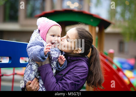 La famiglia felice: la madre e il bambino - bambina camminando in autunno parco: mammy kissing figlia in posa in un parco giochi per bambini vicino fino Foto Stock