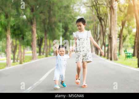 Asian sorella tenere le mani con i bambini piccoli a camminare sulla strada nel parco con raggi di sole. Foto Stock