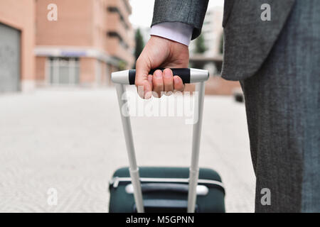 Primo piano di un giovane imprenditore caucasico in un elegante abito grigio tirando il carrello per la maniglia sulla strada Foto Stock