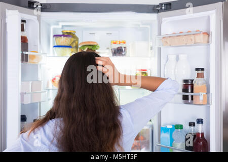 Vista posteriore di un confuso giovane donna cercando in frigorifero aperto Foto Stock