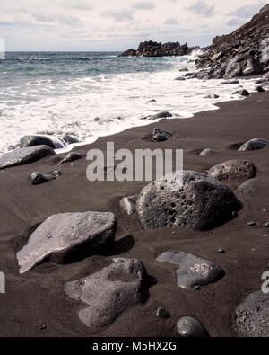 Salinas de Fuencaliente, La Palma. Isole Canarie Spagna. Onde che si infrangono sulla spiaggia di lava. Alcune grandi rocce laviche in primo piano per interesse. Questo piccolo Fuencaliente bay è tipico dell'isola la spettacolare costa. Le rocce laviche salire bruscamente a formare una scogliera. Il sole splende rendendo la schiuma di mare molto bianco e il cielo blu pallido. Le grandi rocce laviche in primo piano sono state erose dal mare che mostra la durezza dell'ambiente marino. Finalmente il mare si erodono le rocce di grandi dimensioni in multa sabbia lavica come il resto della spiaggia. Fotografato con una Ricoh GRII Foto Stock