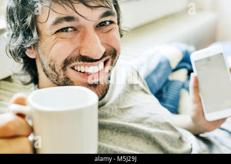 Ritratto di ridere giovane uomo disteso sul letto con lo smartphone e la tazza di caffè Foto Stock