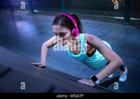 Giovane Donna con cuffie rosa fa pushups nel moderno impianto urbanistico di notte Foto Stock