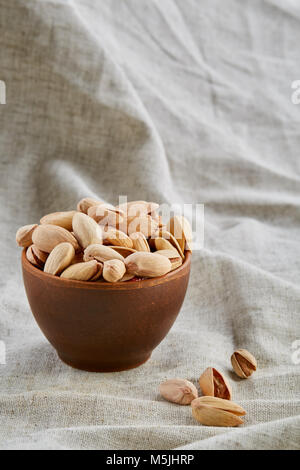 Composizione di materie non pelati pistacchi in argilla vaso in ceramica su grigio chiaro tovaglia in cotone o tovagliolo, qualche spazio copia, closeup, il fuoco selettivo. Guarire Foto Stock