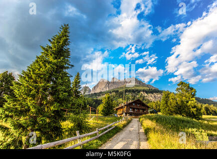 Vista sulle montagne del gruppo del Catinaccio (Rosengarten) con prati e abeti, una strada e di una baita di montagna al di sotto di un blu cielo nuvoloso, Dolomiti, Foto Stock