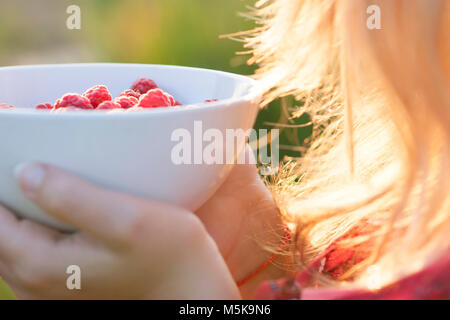 Giovane ragazza con una piastra di lamponi, seduti su erba verde, estate, dessert, sera sunset Foto Stock