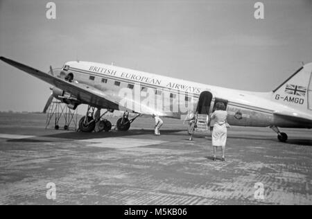 Un Douglas DC-3 Dakota, o C-47, G-AMGD, appartenenti a BEA, British European Airways, presso un aeroporto sconosciuto nel 1960. Un hostess di aria può essere visto a piedi dall'aereo, che ha la porta posteriore aperta, mentre un ingegnere è al di sotto del parafango, e una donna scatta una fotografia del velivolo. Foto Stock