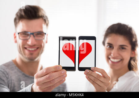 Sorridente Coppia giovane che mostra la forma di cuore unendo il loro telefono cellulare Foto Stock