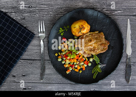 Carne di maiale alla griglia con verdure alla griglia e patate al forno su una piastra nera Foto Stock