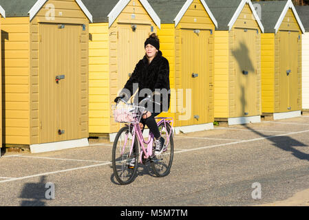 Donna in bicicletta mentre indossa un cappotto e un cappello di pelliccia al sole invernale, davanti a capanne gialle sulla passeggiata, Bournemouth, Dorset, Regno Unito, febbraio. Foto Stock