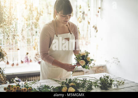 Giovani femmine fioraio mettendo insieme un mazzo di rose mentre si lavora a un tavolo nella sua officina di fiori Foto Stock