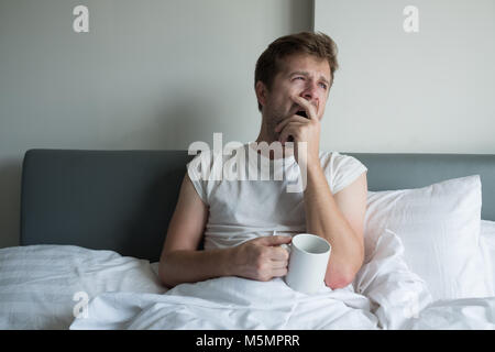 Giovane uomo bello sbadigliare e avente il caffè del mattino a letto Foto Stock