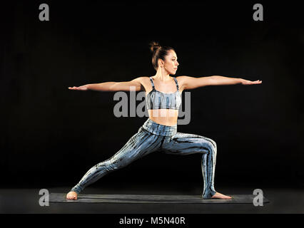 Montare athletic giovane donna dimostrando il guerriero 2 pone in yoga stretching affondo in avanti dritto con le braccia tese su nero Foto Stock