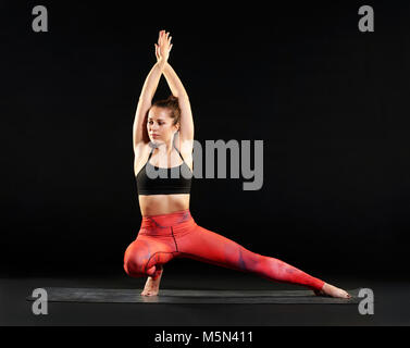 Montare la donna facendo un affondo laterale pongono nello yoga che si estende verso il lato con la gamba estesa e bracci sollevati in una vista frontale sul nero con spazio di copia Foto Stock