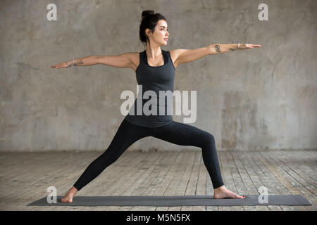 Giovane donna a praticare yoga, facendo Virabhadrasana 2 Esercizio, guerriero due pongono, lavorando fuori, di indossare abbigliamento sportivo, pantaloni neri e top, interni piena leng Foto Stock