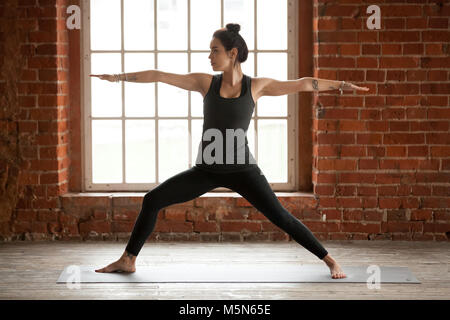 Giovane donna sportiva la pratica dello yoga, facendo Virabhadrasana 2 Esercizio, guerriero due pongono, lavorando fuori, di indossare abbigliamento sportivo, pantaloni neri e top, interni fu Foto Stock