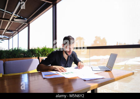 Lavoratore di ufficio ordinamento di carte sul tavolo vicino al computer portatile. Foto Stock