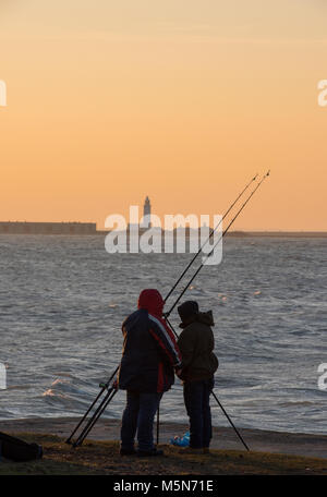 Due pescatori su una spiaggia al tramonto con hurst castle il faro in distanza al tramonto. Bagliore arancione nel cielo di pescatori di mare con aste stagliano sky. Foto Stock