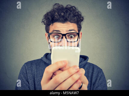 Giovane uomo barbuto in bicchieri usando il telefono con espressione di grande stupore. Foto Stock