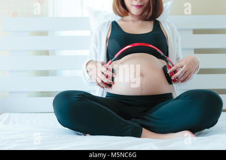 Felice Asian incinta Donna seduta sul letto ascoltare musica con le cuffie Foto Stock