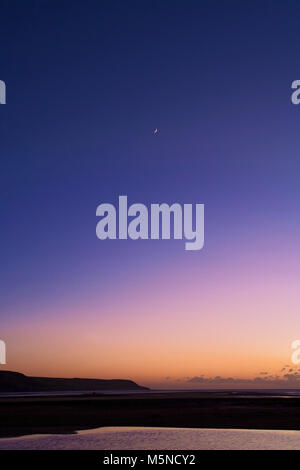 La luna nuova aggiunge un punto focale per un tramonto sulla baia di Barmouth, Galles. Foto Stock