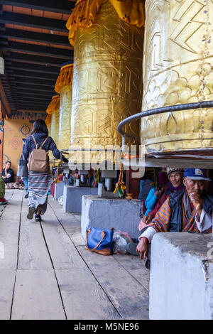 Thimphu, Bhutan. Giovane donna girando le ruote di preghiera presso il National Memorial Chorten mentre altri sedersi sotto. Foto Stock