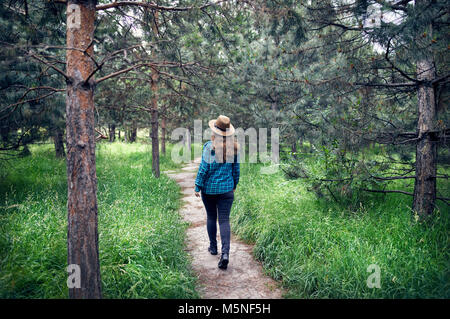 Giovane donna in marrone cappello e verde maglietta controllati con capelli lunghi passeggiate nella foresta di alberi di pino Foto Stock