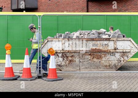 Builder in un elmetto e alta viz camicia utilizzando un telefono cellulare appoggiato su di un saltare riempito di macerie England Regno Unito Foto Stock