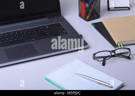 Close-up di comodo posto di lavoro in un ufficio con computer portatile, un mouse, notebook, bicchieri, penna e altre apparecchiature posa sulla tavola su mobili di sfocatura dello sfondo. Foto Stock